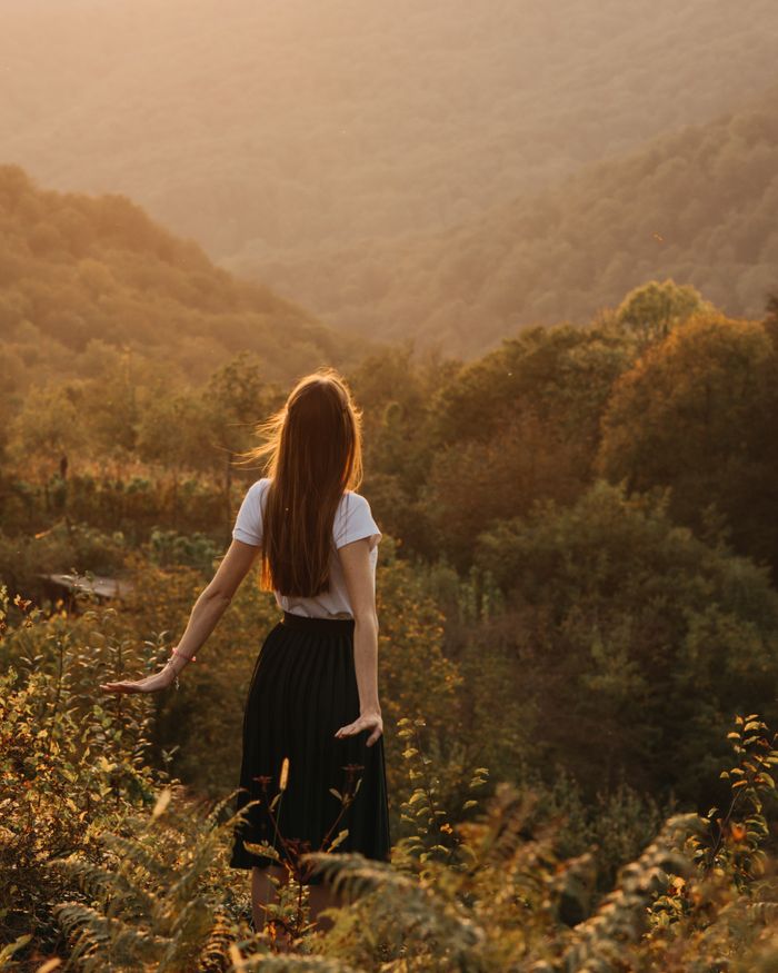Back view of unrecognizable young female in white shirt and black skirt spreading arms back while standing on picturesque hill with lush green trees and plants and looking away in evening sunlight