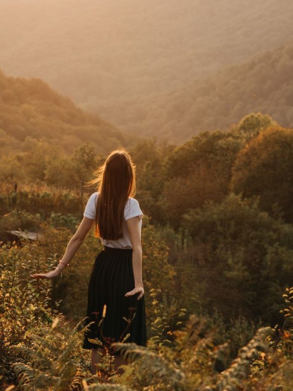 Back view of unrecognizable young female in white shirt and black skirt spreading arms back while standing on picturesque hill with lush green trees and plants and looking away in evening sunlight